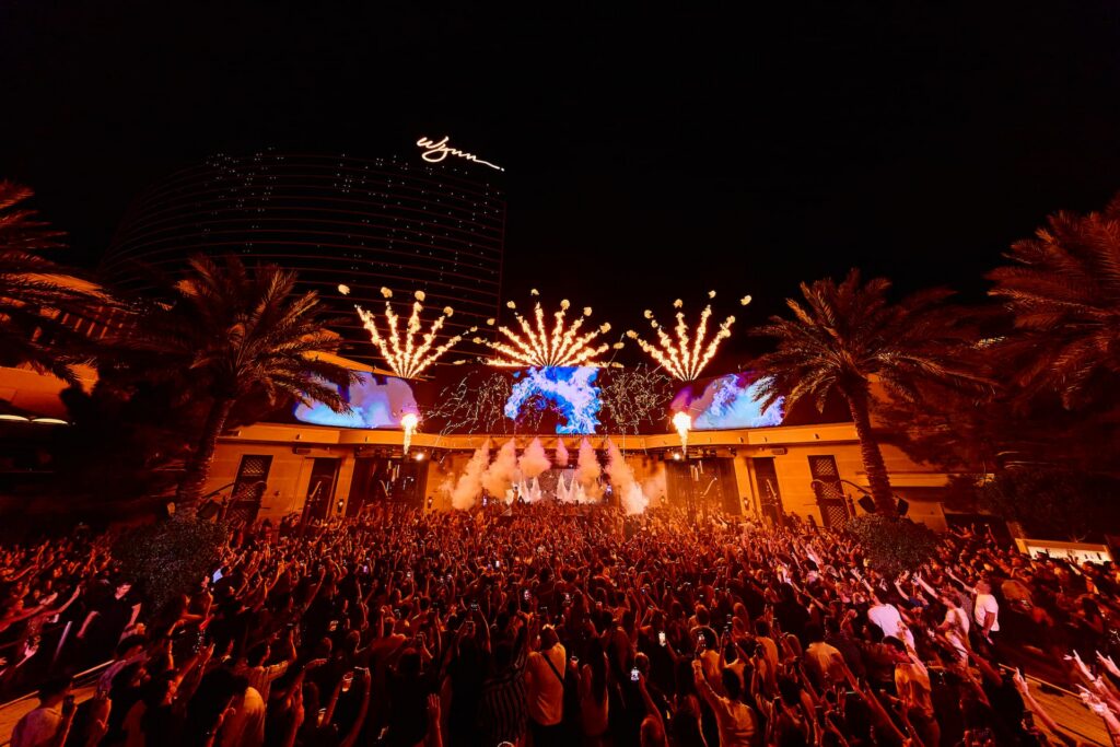 Large nighttime crowd at an outdoor XS Nightclub event with stage lights, pyrotechnics, palm trees, and Wynn Las Vegas in the background.